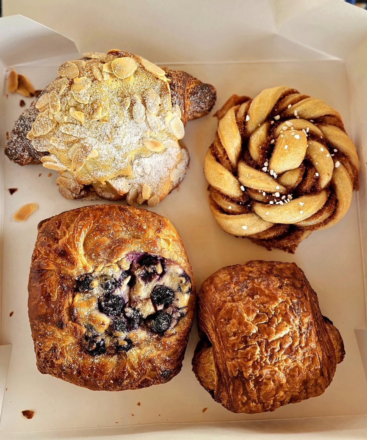 Four pastries in an open box seen from above: an almond croissant dusted with sugar, a cardamom knot with pearl sugar, a blueberry danish and a pain au chocolat.